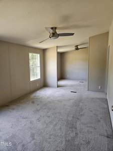 An empty room with beige walls and carpeting, featuring two ceiling fans and large windows letting in natural light. The room is long and narrow, with modern fixtures and minimal decor.