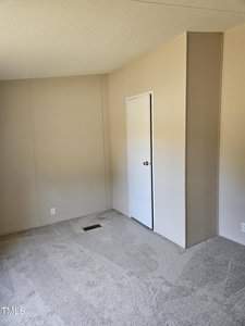 An empty, beige-colored room with carpeted flooring, a small closet with a white door, and a vent on the floor. The walls are plain and the room is well-lit.