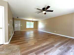 A spacious, empty living room with light wood flooring, beige walls, and a ceiling fan. A large window and a small hanging light fixture brighten the room. The entrance door is visible in the background.