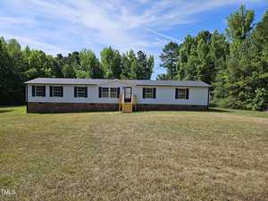 A single-story house at 2142 Mountain Creek Road features a dark roof and light-colored siding, enveloped by a grassy lawn and tall green trees. The Oxford blue sky is peppered with clouds, and a small wooden porch with steps leads to the inviting entrance.
