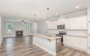 Modern open-concept kitchen and living area with white cabinets, stainless steel appliances, a large island with granite countertops, pendant lighting, and wood flooring. A fireplace is visible in the adjacent living space.