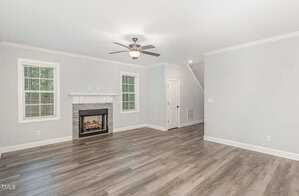 A bright, empty living room with light gray walls and wood flooring. It features a ceiling fan, a white-trimmed gas fireplace with a stone surround, two windows, and a closed door. A staircase is visible on the right.