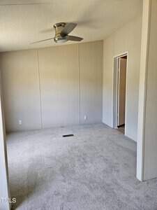 An empty room with light beige walls, a gray carpet, and a ceiling fan. The left wall features a window not visible in the image. There is a doorway to the right leading to another room. The space is bright and unfurnished.
