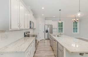 A modern kitchen featuring white cabinetry, granite countertops, and stainless steel appliances, including a refrigerator and oven. A large island with a sink is centered, and pendant lights hang from the ceiling. The room has light wood flooring.