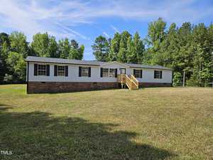 A single-story white house with dark shutters and a brick foundation is situated in a spacious grassy yard. A wooden staircase leads to the front entrance. Tall trees surround the property under a clear blue sky.