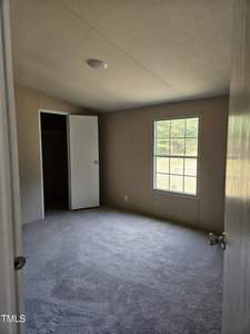 A small, empty room with beige walls and a carpeted floor. There's a closed window on one wall, and an open door leading to a closet. Light enters through the window, illuminating the space.