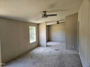 A spacious, empty room with beige walls and carpeted flooring. Two ceiling fans are installed, and sunlight streams through a window on the left, illuminating the space.
