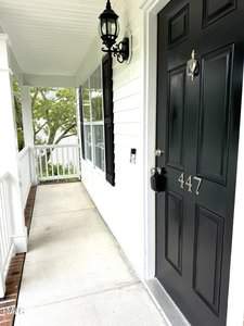 A black front door with the number 447, featuring a silver knocker and handle. The door is set in a white-paneled house with a porch, white railing, and a black lantern light fixture above. There are green trees visible in the background.