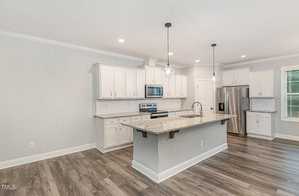 Modern kitchen with white cabinets, stainless steel appliances, and a large central island with a granite countertop. Two pendant lights hang above the island. Wooden flooring adds warmth, and a window offers natural light.