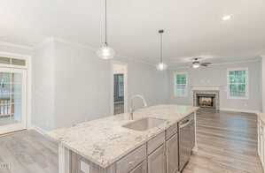 Modern kitchen with a large granite island featuring a sink and pendant lighting. Open layout leads to a living room with a fireplace, ceiling fan, and windows. Light wood flooring and a door to a covered patio enhance the space.