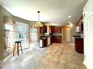 Bright kitchen with tiled floors, wooden cabinets, and floral curtains. Features a central light fixture, a stool by the windowed door, and stainless steel appliances. Ample natural light fills the room.