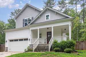 A modern two-story house with gray siding and white trim features a covered porch, a dark red front door, and a double garage. It is surrounded by lush green grass and trees, creating a peaceful, natural setting.