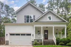 A two-story house with light gray siding and white trim, featuring a covered porch. It has a two-car garage with a paneled door. Surrounded by trees, the home has a neatly landscaped front yard with green shrubs.