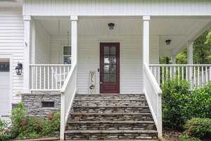 Front porch of a house with a red door, white siding, and a stone staircase. A vertical "Welcome" sign stands by the door. Lush greenery surrounds the porch, and a hanging light fixture is visible by the door.