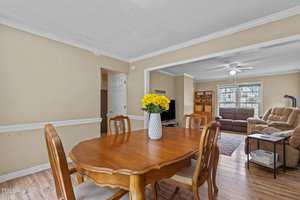 Dining room with a wooden table and chairs, decorated with a vase of yellow flowers. Beyond, a cozy living area features a brown recliner, a large window, and a TV on a wooden stand. Walls are light-colored, and hardwood floors are present throughout.