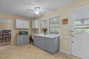 A cozy kitchen with light yellow walls, white upper cabinets, and blue lower cabinets. A white ceiling fan and a bouquet of red flowers are on the counter. A door with a window leads outside, and a microwave sits on the counter.