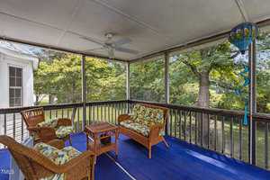 A screened porch with wicker furniture featuring floral cushions on a blue rug. A small table is in the center, and a ceiling fan is above. A decorative wind chime hangs on the right. Lush greenery is visible through the screens.