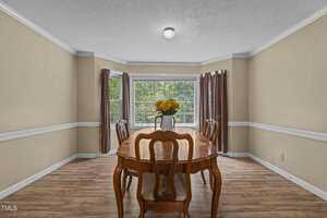A dining room with a wooden table and four chairs. A vase of yellow flowers is centered on the table. The room has beige walls, a large bay window with brown curtains, and wooden flooring. Natural light fills the space.