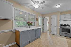 A kitchen with light-colored walls and beige tile flooring features a stainless steel stove, white cabinets, and a ceiling fan. A vase with red roses sits on the white countertop below a window with blinds. Doorways are visible in the background.