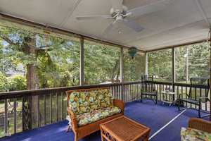 A cozy screened porch with a ceiling fan, adorned with wicker furniture featuring floral cushions. A blue carpet covers the floor, and trees surround the area outside the screened windows. A heart-shaped wind chime hangs from the ceiling.