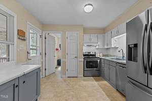 A kitchen with light-colored walls and tile flooring. It features stainless steel appliances, including a refrigerator and oven. White cabinets line the walls, and there is a window above the sink with blinds, offering a view outside.