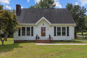 Nestled at 103 Meadow Brook Lane, this quaint white house in Oxford boasts a dark roof and brick chimney, with a charming porch welcoming you through its single door flanked by two windows. It's embraced by a green lawn and trees under the expansive clear blue sky.