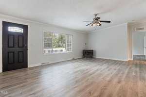 A spacious living room with light wood flooring, neutral-colored walls, a dark wooden front door, and a large window letting in natural light. A ceiling fan with lights is centered on the ceiling, and a small fireplace is in the corner.