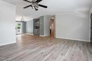 Spacious living area with light wood flooring and gray walls. Ceiling fan with black blades above. In the background, there's an open kitchen with gray cabinets and a chandelier, leading to a deck via glass doors. Natural light fills the room.