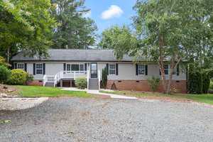 Nestled in the serene setting of Franklinton, this single-story home at 1531 Wayside Farm Road boasts a gray exterior and black roof, surrounded by lush trees and shrubs. A charming white front porch welcomes you, with a gravel driveway and patch of grass under the clear blue sky.