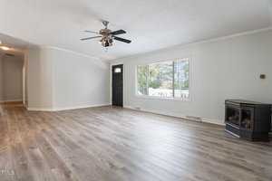 A spacious living room with light-colored wood flooring and white walls. A black ceiling fan hangs from the ceiling. A large window illuminates the room, and a fireplace sits in one corner. A hallway leads to other parts of the house.
