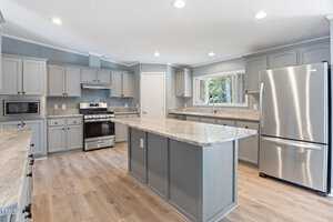 A modern kitchen with gray cabinets, a large island with marble countertops, a stainless steel refrigerator, an oven, and a microwave. The floor is wooden, and natural light comes through a window above the sink.