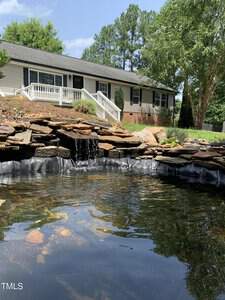 A serene backyard scene with a small waterfall flowing into a pond surrounded by rocks. A single-story house with a porch and railing is visible in the background, framed by green trees and a clear blue sky.