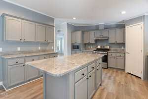 A modern kitchen with gray cabinets, a marble countertop island, stainless steel appliances, and wooden flooring. The space is well-lit with recessed lighting and has a neutral color palette.