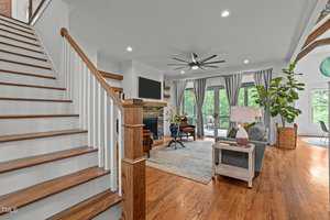 A spacious, elegant living room with hardwood floors and a large staircase. There's a stone fireplace, a modern ceiling fan, and large windows with curtains offering a view of greenery. A cozy seating area with a rug and plants completes the space.