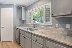 A modern kitchen with light gray cabinets, light granite countertops, and a stainless steel sink under a large window with blinds. The walls are a soft blue-gray, and the floor is light wood. A white door is on the left.