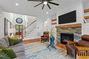 Modern living room with wooden floors, a gray sofa, and two brown leather armchairs. A stone fireplace features a TV above it. A large rug covers part of the floor. Wooden shelves are on the wall, and a ceiling fan hangs from the ceiling. Stairs in the background.