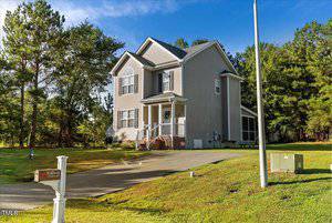 Two-story gray house with a sloped driveway in a suburban area. Surrounded by green trees and grass, the house features a small front porch and multiple windows. A "Welcome" sign is visible on a post near the driveway. Clear blue sky above.