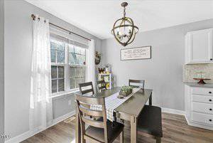 A dining area with a wooden table set for four, featuring a white runner and small centerpiece. A large window with sheer curtains lets in light. On the wall is a "Bless" sign. A modern chandelier hangs above, with white cabinetry in the background.