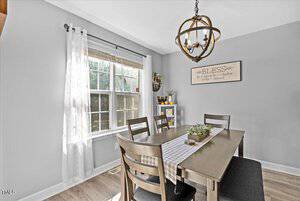 Dining room with a wooden table and four chairs. A modern chandelier hangs above, and a "Bless" sign decorates the gray wall. Light streams in through a window with sheer curtains. A small plant centerpiece and decorative shelf complete the cozy space.