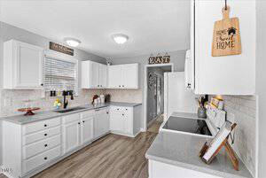 A modern kitchen with white cabinets, gray countertops, and wooden flooring. Decorative signs are on the walls and cabinets. A fruit bowl and a candle are on the countertop. The kitchen leads to a hallway with an "EAT" sign overhead.