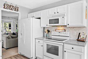 A bright, modern kitchen with white cabinets and appliances. A "EAT" sign is above a doorway leading to a living area with a gray sofa. The backsplash features light tiles, and there's a cookbook on display beside a knife block on the countertop.