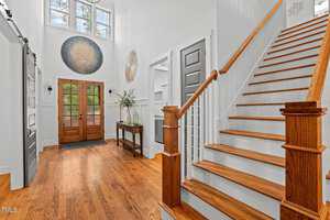 A bright, airy foyer with wooden floors and stairs. It features a large front door with glass panels, decorative wall art, a console table with a plant, and a staircase with white railing. High windows let in natural light.
