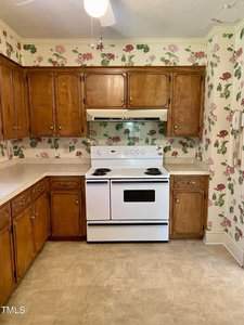 A vintage kitchen features wooden cabinets, a white electric stove with an oven hood, and floral wallpaper with roses. The floor is covered with beige linoleum. A rotary dial wall phone is mounted to the right.