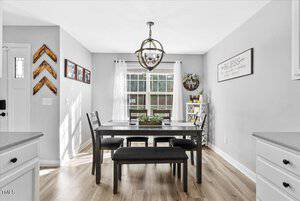 A bright dining room with a dark wood table and chairs, a bench, and a chandelier. A window with sheer curtains lets in natural light. The walls are gray with decorative frames and a sign, while the floor is light wood.