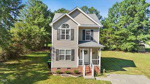 Nestled at 117 Chalet Lane, this two-story house with beige siding and white trim graces a spacious lawn. It boasts a charming front porch with steps, black shutters, and a driveway, surrounded by green trees under the clear blue sky.