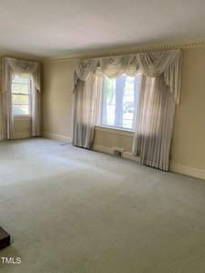 A spacious living room with beige walls and light green carpet. Large windows are adorned with elegant cream drapes, allowing natural light to flood the room.