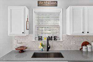 A bright kitchen features white cabinets, a gray countertop, and a tile backsplash. Below a window with blinds is a black faucet and a "HOME" sign. Decorative items and jars sit on the counter, and a wooden sign above reads "The Williams, Est. 2023.