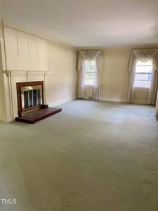 A spacious living room with a fireplace on the left, surrounded by light-colored walls. Two windows with curtains allow natural light to fill the room, which features a pale carpeted floor. The room is unfurnished.