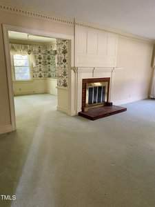 A spacious living room with light-colored carpet and a white, paneled fireplace with a brick base. An adjacent room with floral wallpaper and a window letting in natural light is visible. The walls and molding are painted in neutral tones.