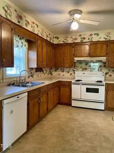 Vintage kitchen with wooden cabinets, floral wallpaper, and a white stove. A ceiling fan with a light hangs above. A dishwasher and window with blinds are near the sink.